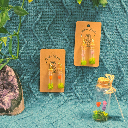 Two sets of earrings on cards with a small decorative bottle and plant on a textured teal background.